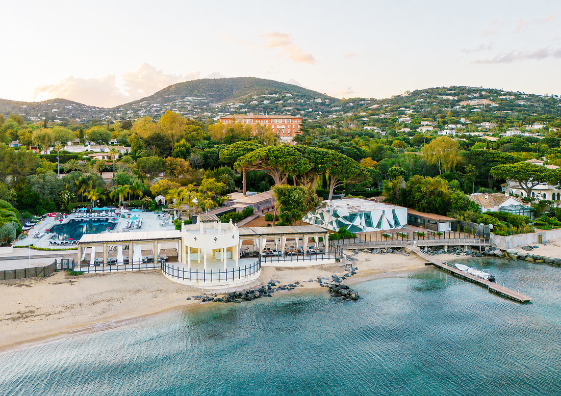 Luftaufnahme einer mediterranen Hotelanlage mit weitläufigem Garten, Pool und Strandpromenade am Meer, umgeben von grünen Hügeln – das COMO Le Beauvallon am Golf von Saint-Tropez.