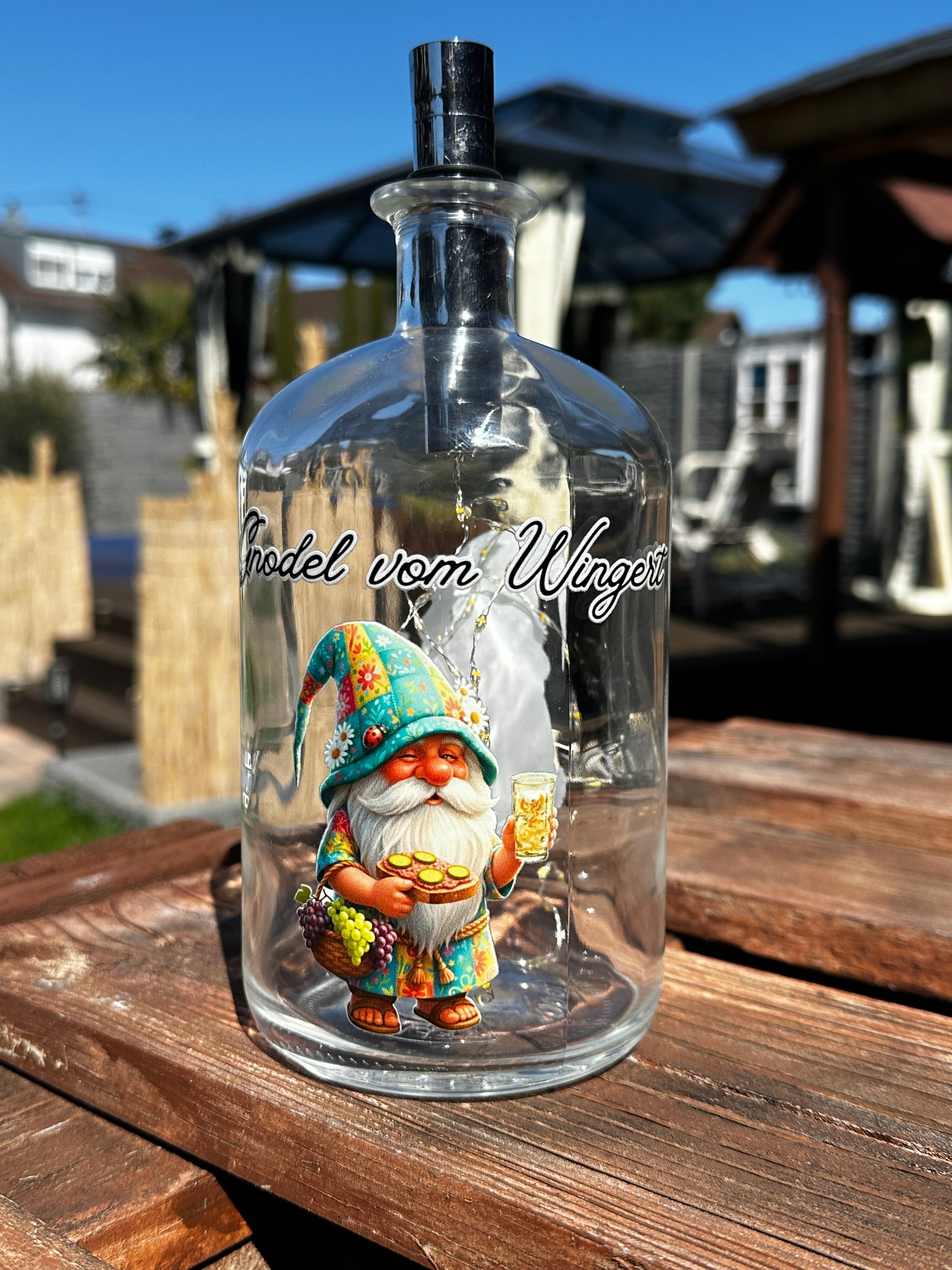 Dekorative Flaschenlampe mit Pfälzer Dubbewichtel Gnodel vom Wingert auf einem rustikalen Holztisch im Garten. Beleuchtetes Glas-Deko-Objekt mit Wichtel-Motiv, Schriftzug und Lichterkette als Geschenkidee für Pfalz-Liebhaber und Gartenbesitzer.
