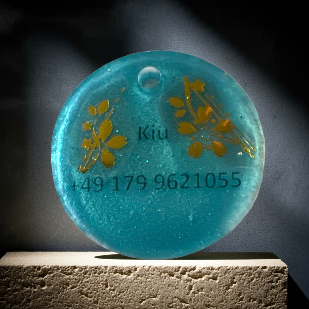 Runder, blau-türkiser Tier-Anhänger mit eingearbeiteten Gold-Elementen, der auf einem beleuchteten Sockel präsentiert wird.