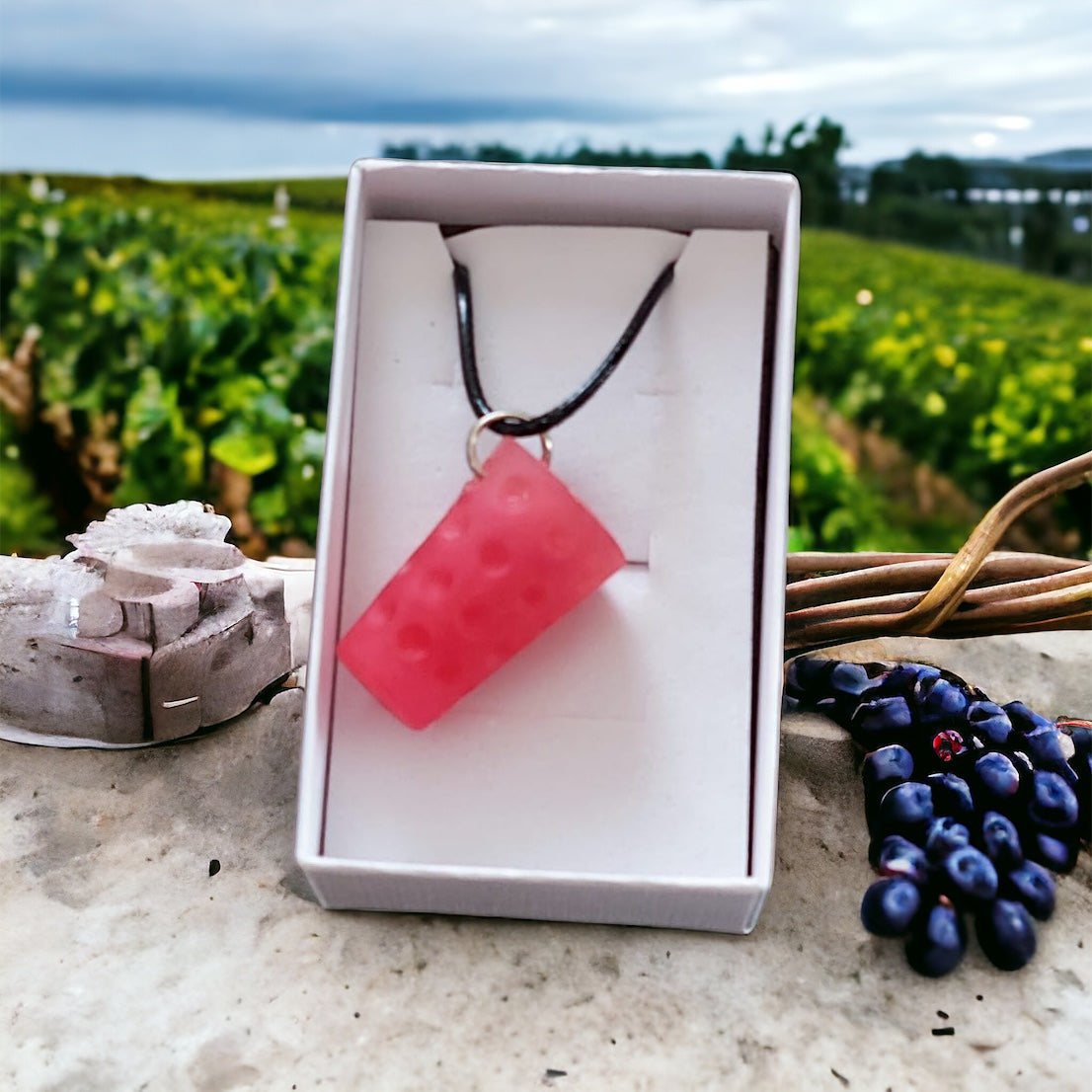 Pinkfarbener Dubbeglas-Anhänger in einer Geschenkbox, fotografiert in der Natur vor Weinreben.