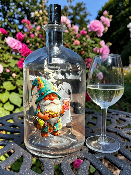 Pfälzer Flaschenlampe mit Dubbewichtel Gnodel vom Wingert – stimmungsvolle Gartenbeleuchtung und Geschenkidee für Pfalz-Fans. Beleuchtete Glasflasche mit Wichtel-Motiv, Dubbeglas und Weintrauben auf einem Gartentisch neben einem Glas Weißwein vor Rosenbüschen.