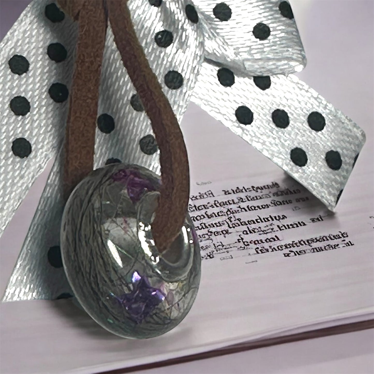 Runder Gedenkschmuck-Charm mit eingearbeiteten violetten Elementen, präsentiert auf einem weiß-schwarz gepunkteten Band vor einem Schriftstück.