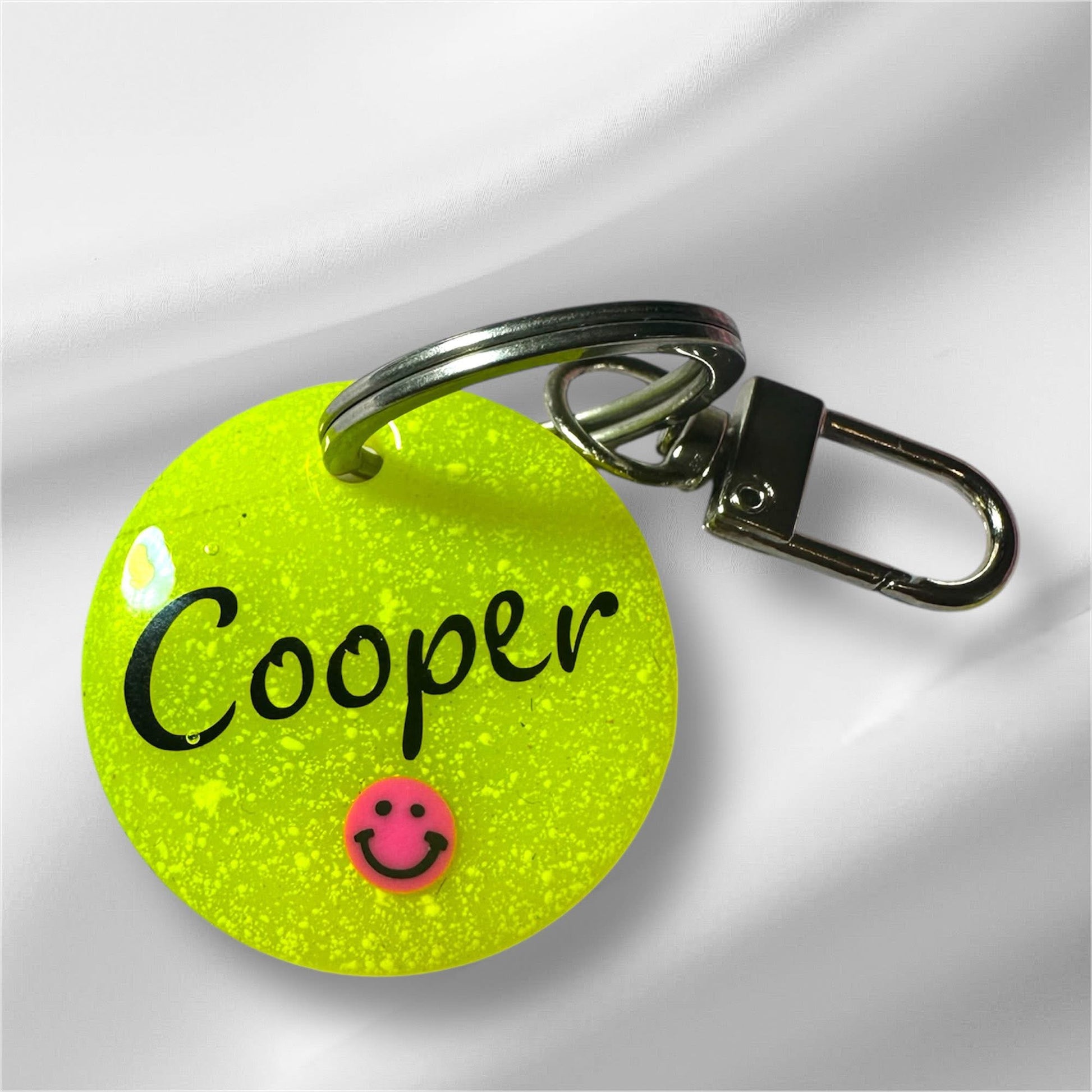 Leuchtend gelber, runder Tier-Anhänger mit dem Namen „Cooper“ und einem kleinen Smiley-Symbol