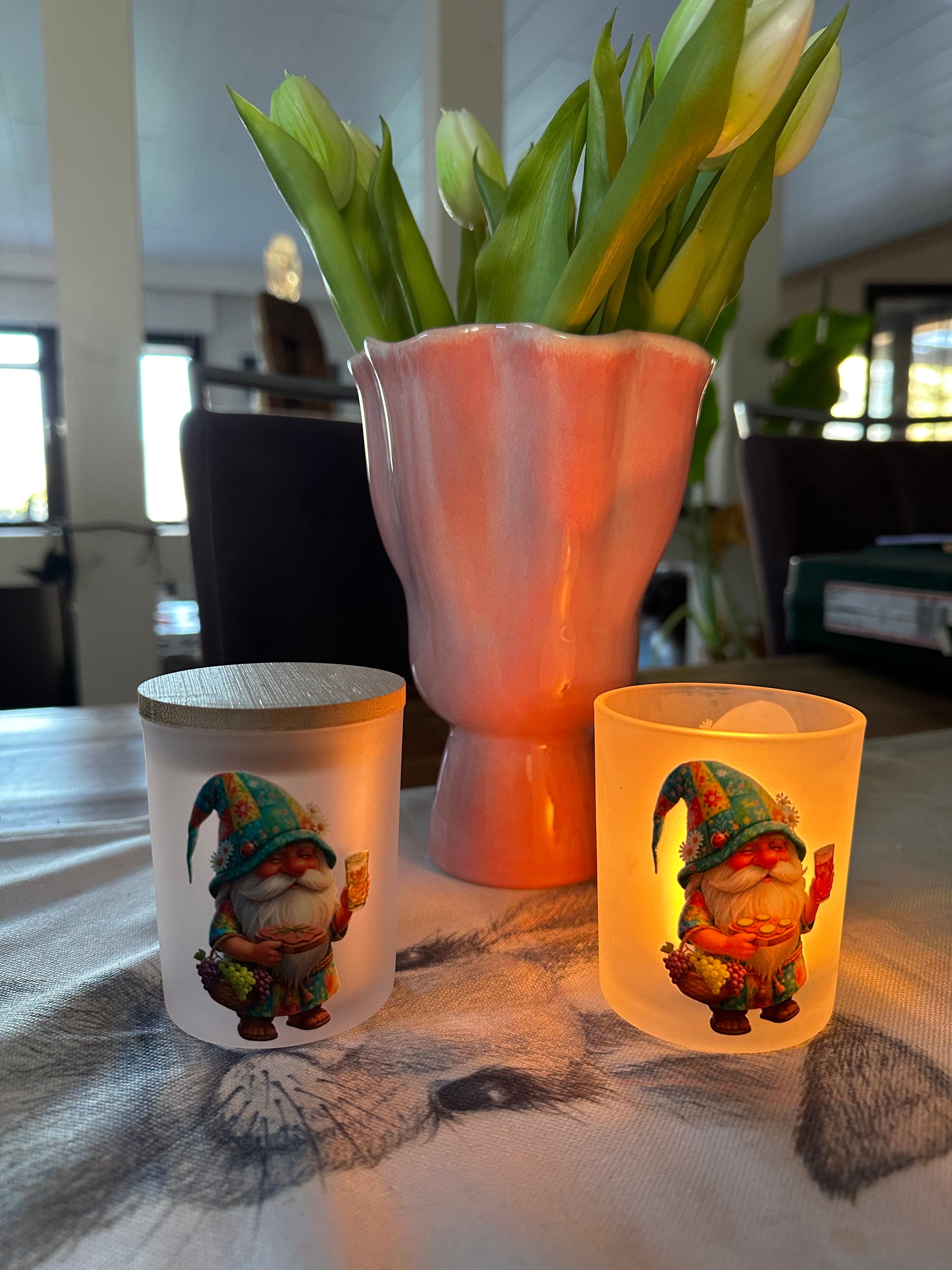Zwei gefrostete Windlicht-Gläser mit Pfälzer Wichtel-Motiv auf einem Holztisch neben einer rosa Blumenvase mit weißen Tulpen. Ein Glas ist mit einem Holzdeckel verschlossen, das andere wird von einem Teelicht von innen beleuchtet und zeigt den Wichtel mit einer Schorle.