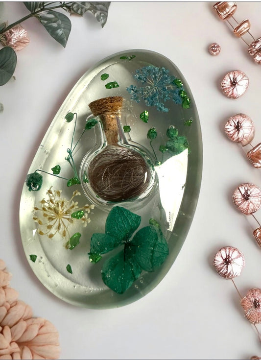 Ovaler Handschmeichler aus klarem Epoxidharz mit einer eingearbeiteten kleinen Glasflasche, Phiole für Tierhaar-Andenken, grünen Trockenblumen und Farn-Elementen auf rosafarbenem Hintergrund.