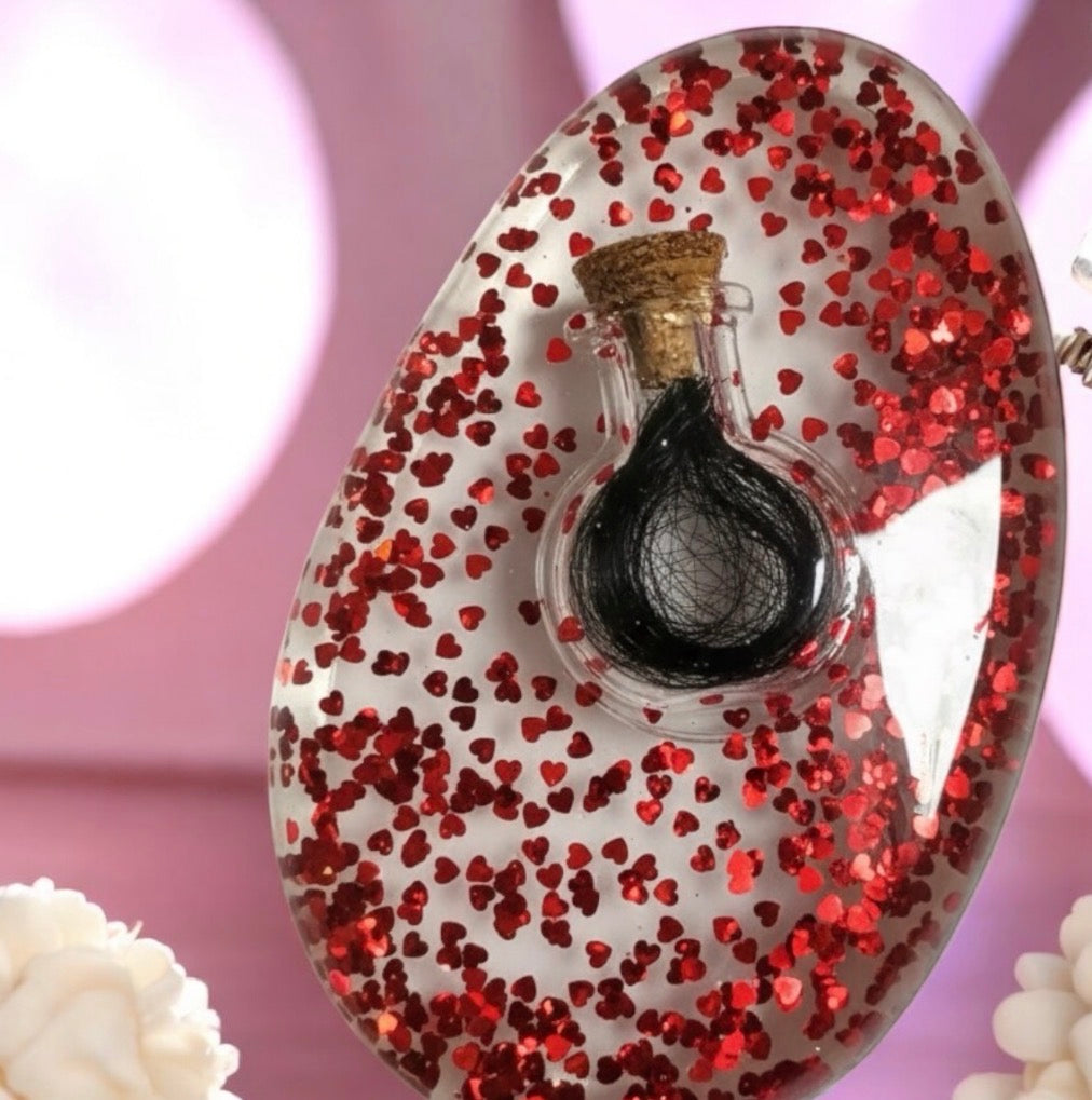 Ovaler Handschmeichler aus klarem Epoxidharz mit einer eingearbeiteten kleinen Glasflasche für Tierhaar-Andenken, gefüllt mit zahlreichen roten Glitzer-Herzen.