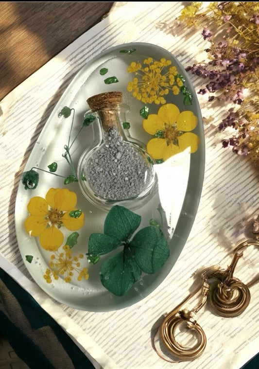 Ovale Gedenkplatte aus transparentem Epoxidharz mit einer eingearbeiteten kleinen Glasflasche für Asche, gelben und grünen Trockenblumen sowie dekorativen Goldelementen.