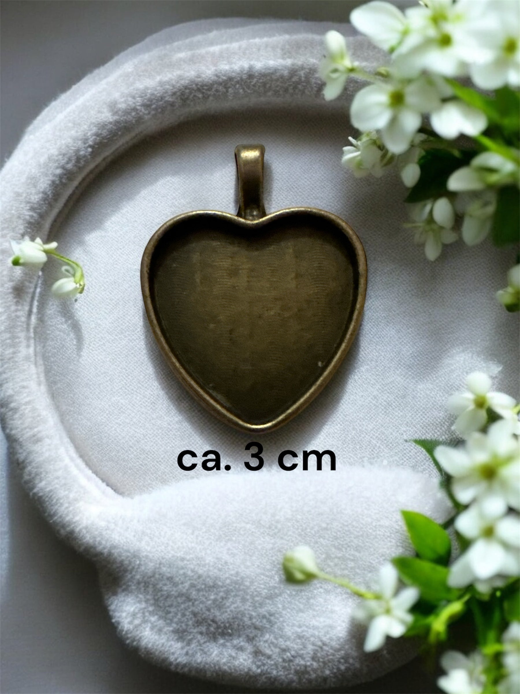 Nahaufnahme einer herzförmigen Gedenkschmuck Fassung in Silber-Optik mit ca. 3,5 cm Breite auf einem floralen Hintergrund.