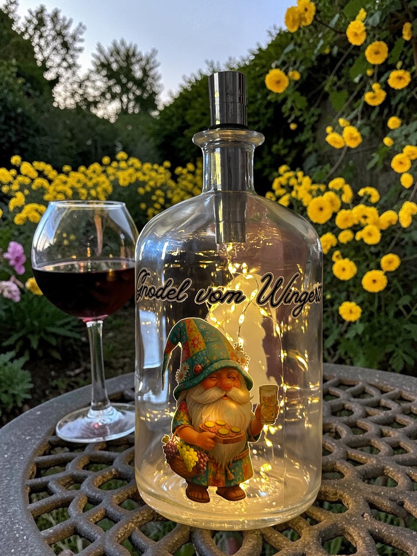 Beleuchtete Weinflaschenlampe mit Dubbewichtel Gnodel vom Wingert bei Dämmerung im Garten. Gemütliche Abend-Atmosphäre mit warmweißem LED-Licht in der Flasche, einem Glas Rotwein und gelben Blumen im Hintergrund – ideales Pfälzer Geschenk.