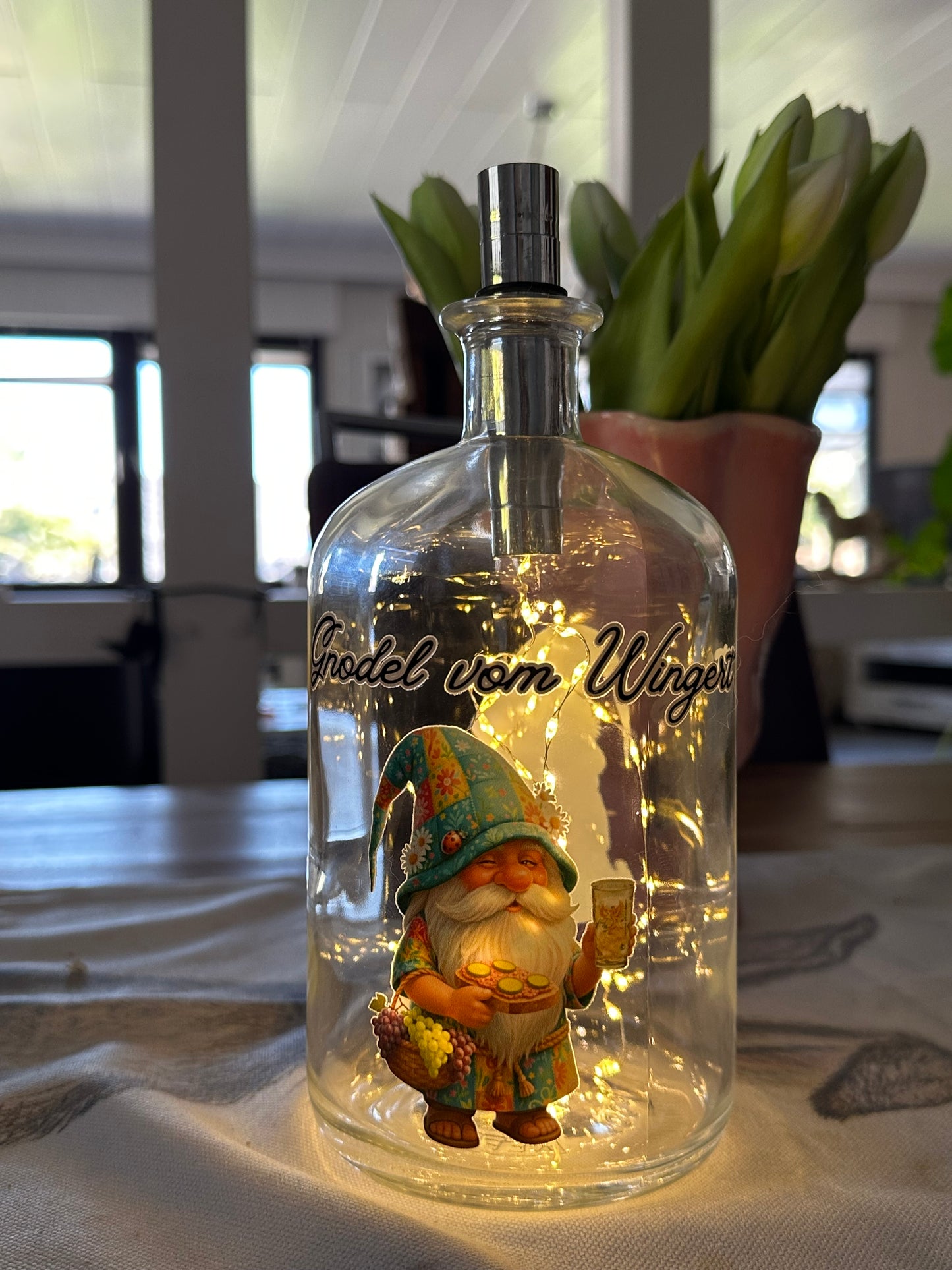 Pfälzer Flaschenlampe „Gnodel vom Wingert“ als stimmungsvolle Innenbeleuchtung auf einem gedeckten Tisch. Die beleuchtete Glasflasche mit Dubbewichtel-Motiv steht neben einer Vase mit hellen Tulpen und sorgt für ein gemütliches Ambiente im Haus.