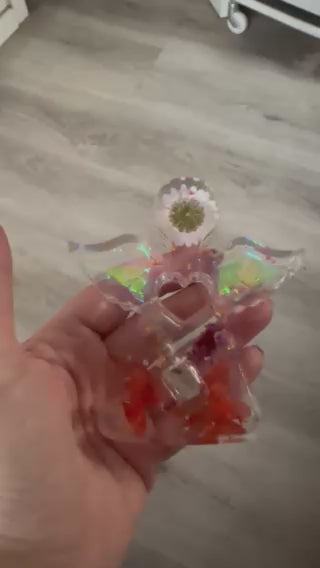 Video-Nahaufnahme einer Gedenk-Figur in Engelform aus klarem Epoxidharz, die in der Hand gehalten wird. Im Inneren sind bunte Trockenblumen und eine kleine Glasphiole für Tierhaar-Erinnerungen sichtbar.