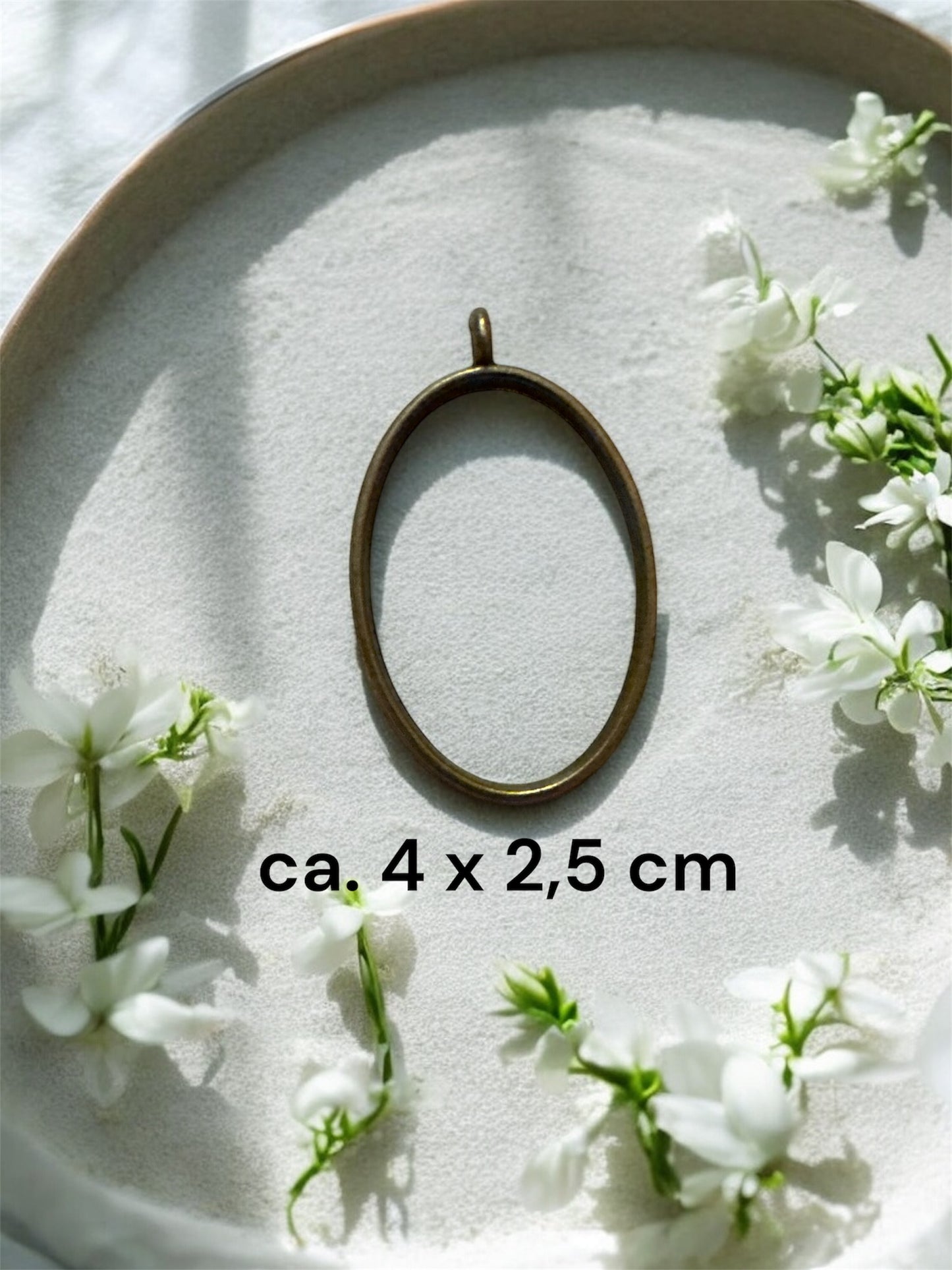 Nahaufnahme einer ovalen Gedenkschmuck Fassung aus Messing mit ca. 4 x 2,5 cm auf hellem Untergrund mit weißen Blüten.