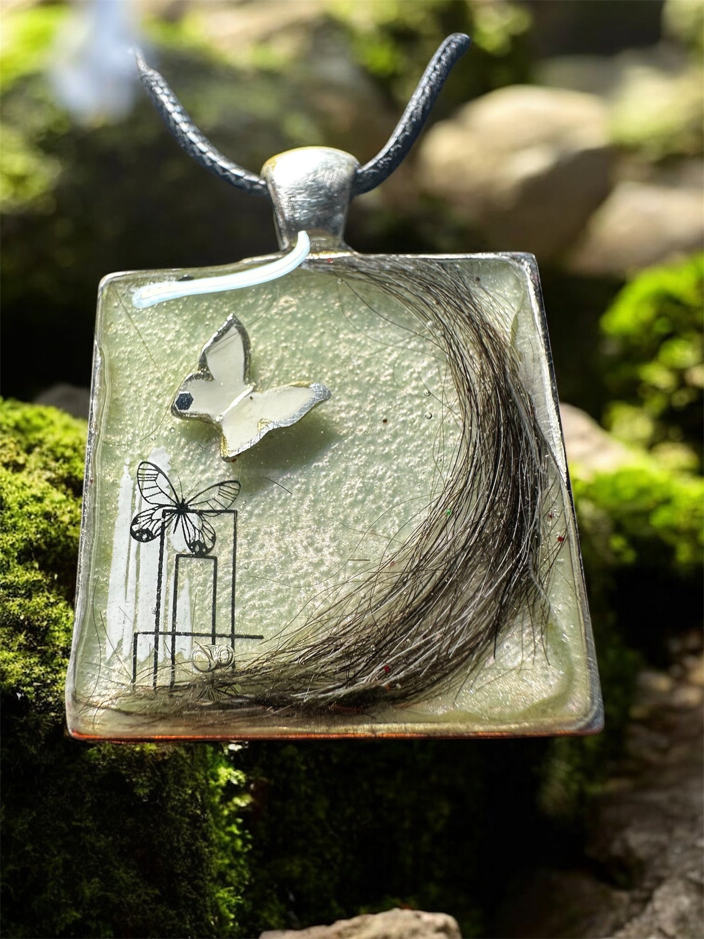 Quadratischer Gedenkschmuck Anhänger mit einer eingearbeiteten dunklen Tierhaarsträhne auf hellem Untergrund.