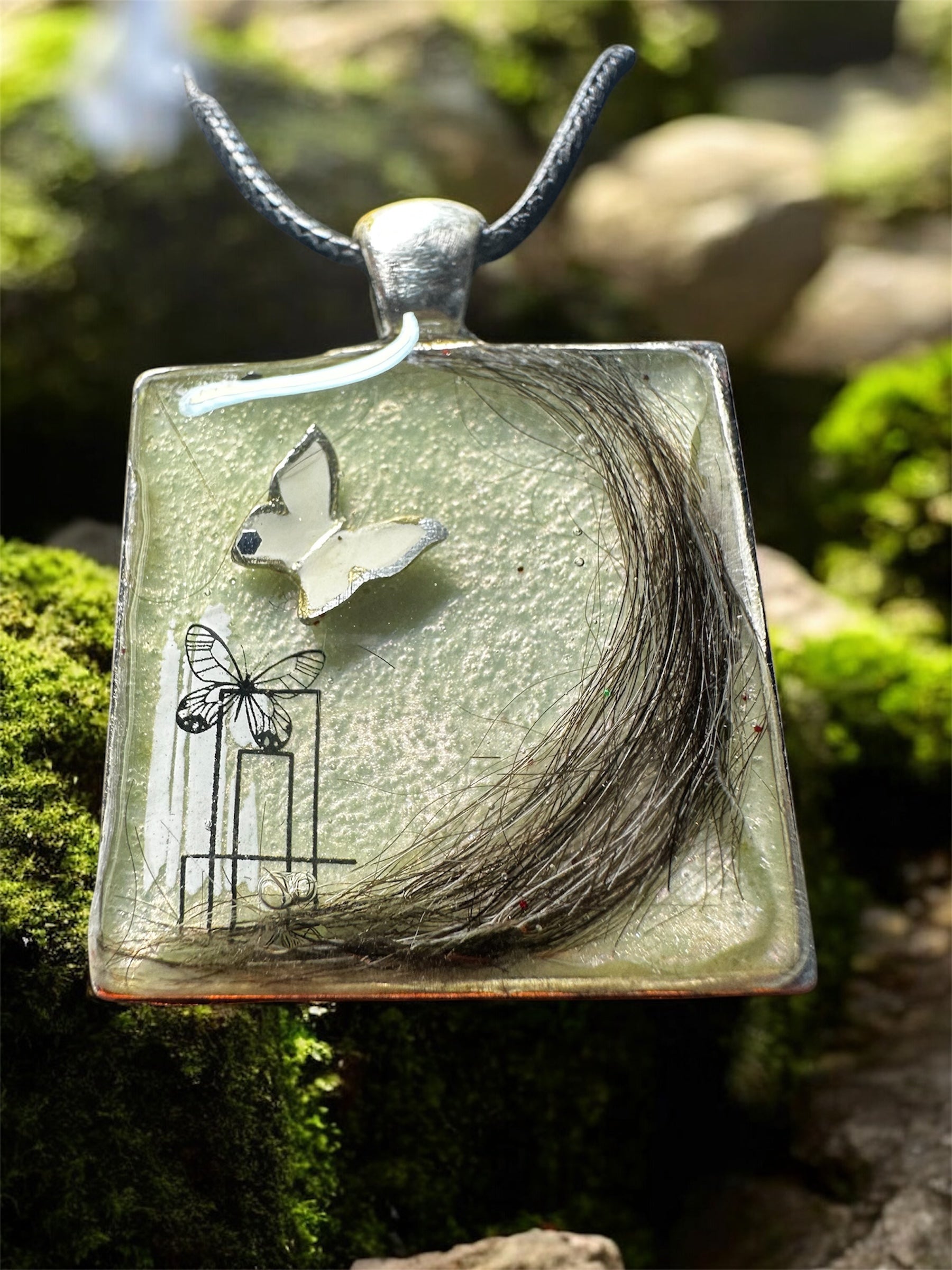 Quadratischer Gedenkschmuck Anhänger mit einer eingearbeiteten dunklen Tierhaarsträhne auf hellem Untergrund.