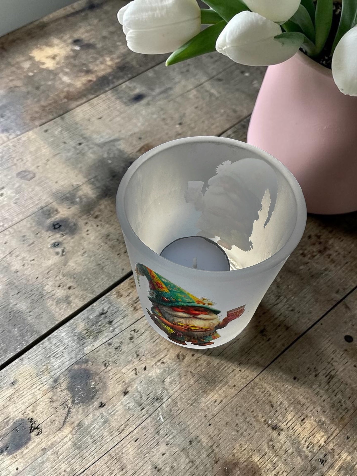 Blick von oben in das leere Gnodel-Windlicht aus gefrostetem Glas. Man sieht das Teelicht am Boden und die Transparenz des Pfälzer Wichtel-Motivs auf der satinierter Glaswand. Das Glas steht auf einem rustikalen Holztisch neben einer rosa Vase.
